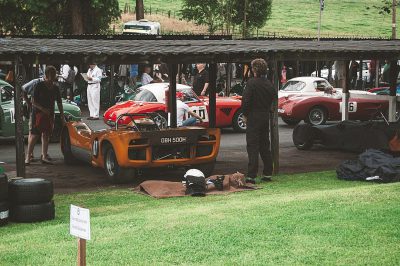 A photo of the garages at an old English racing festival, with classic race cars and crew members working on their vehicles in front, shot from behind as if taken in the style of someone standing near the grassy ground looking up towards them. The scene is set against a backdrop of greenery and traditional wooden structures. In one corner stand two men dressed like film stars working on a cylindrical orange Le Mans car with black wheels and the white numbers "08" written on it, chatting while they work on another red sports motorcycle.