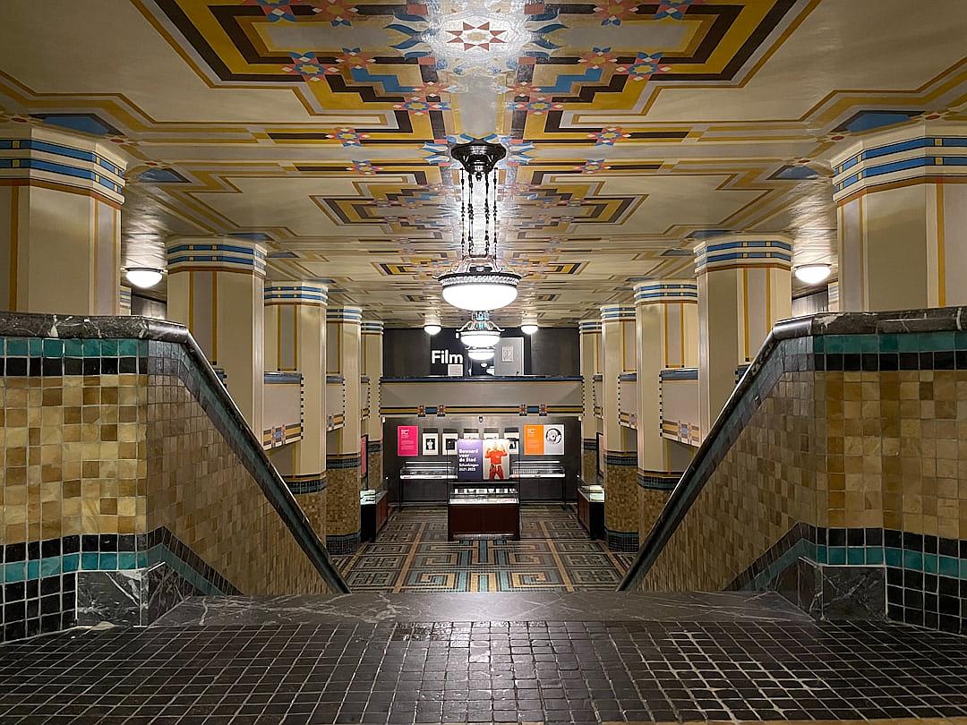 A photograph of the lobby interior showing two marble staircases with dark brown and light blue tiles, the walls adorned in geometric patterns, above it is a ceiling painted yellow, black and red in the style of Art Deco, above that there hangs another chandelier in the style of Art Deco hanging from one side, at ground level on either side are glass doors leading to different areas such as movie screens and posters. In front of each door stands an elegant reception desk made of rosewood. The overall atmosphere feels luxurious and glamorous.