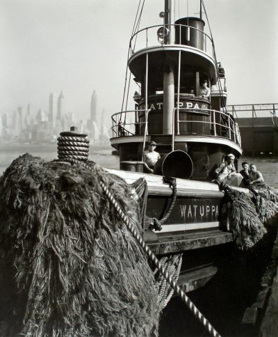 black and white photo of the stern of an old small city harbor ship with a thick pile of dark brown, fluffy wet straw on deck. The name "WATUT tocca" is written in black letters on its side. People sit around it as if they were sitting inside their boat. In the background we see the New York City skyline.