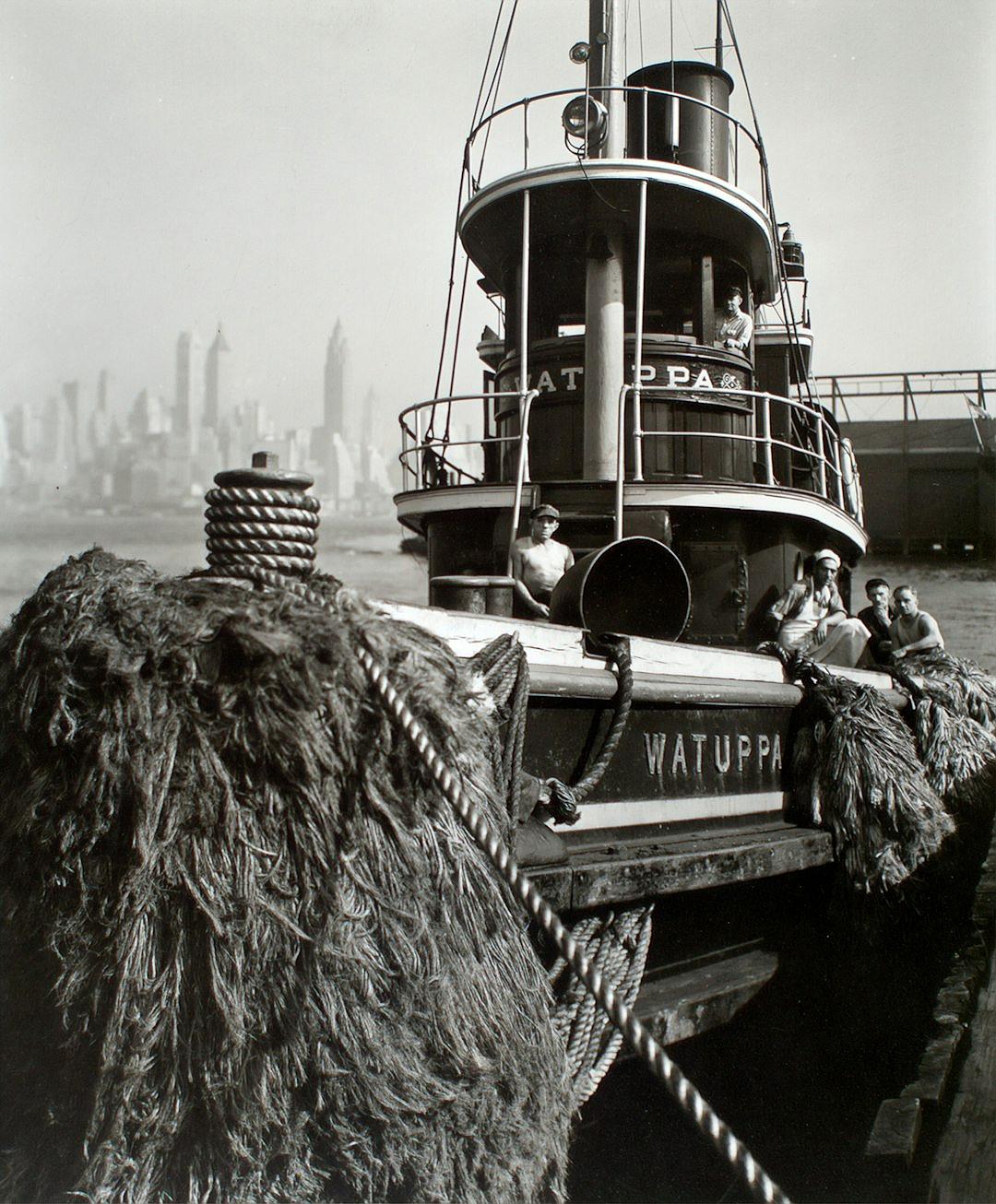 black and white photo of the stern of an old small city harbor ship with a thick pile of dark brown, fluffy wet straw on deck. The name “WATUT tocca” is written in black letters on its side. People sit around it as if they were sitting inside their boat. In the background we see the New York City skyline.