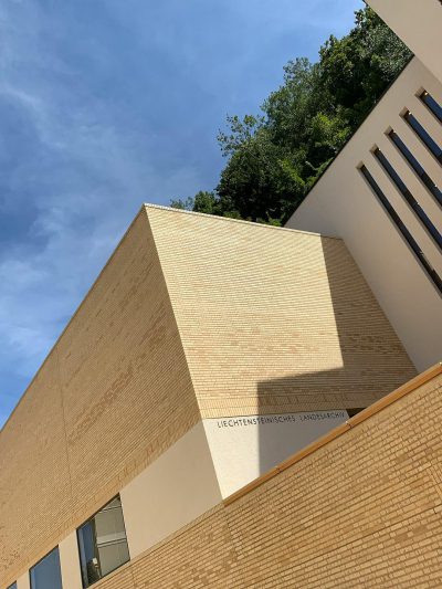 Photo of the exterior wall, modern architecture in the style of [Tadao Ando](https://goo.gl/search?artist%20Tadao%20Ando), beige brick and concrete with white metal details. View from below. The building is located on top of a hill overlooking trees and a blue sky. There is "LFaculty" written as a logo on one side of the facade, "landscape format magazine".