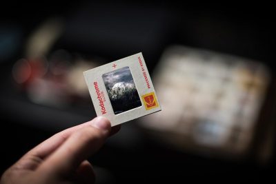 A hand is holding an old photograph with the KODAK logo showing mountains and clouds in the background. The photo is on top of another smaller polaroid card that says "Kodak" in red letters. In front there is a blurred out foreground with an out of focus black background. The Polaroids are placed inside a plastic sleeve for protection.