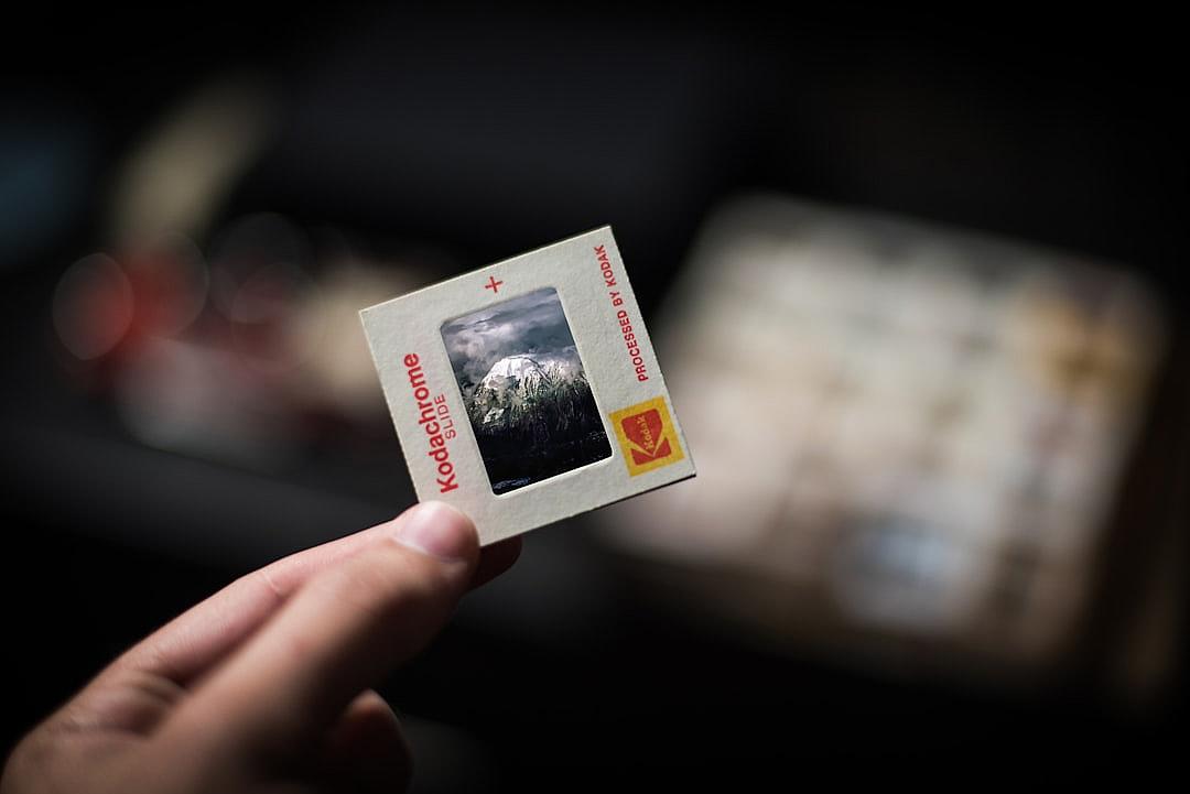 A hand is holding an old photograph with the KODAK logo showing mountains and clouds in the background. The photo is on top of another smaller polaroid card that says “Kodak” in red letters. In front there is a blurred out foreground with an out of focus black background. The Polaroids are placed inside a plastic sleeve for protection.