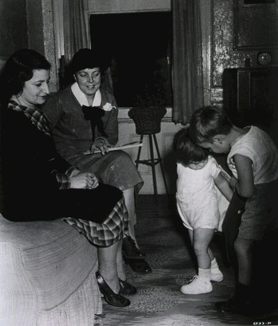 An old black and white photo shows two women sitting on a sofa, one wearing a plaid skirt and shoes while looking at another woman standing in front with her hands folded across her chest. She wears a plain short sleeve top, long pants, and child-like ankle socks. They look down at a small girl playing near them. The room feels old fashioned and dark but there is light coming from the window. The little girl's face peeks out behind a table. The mother looks happy, perhaps just having seen it.