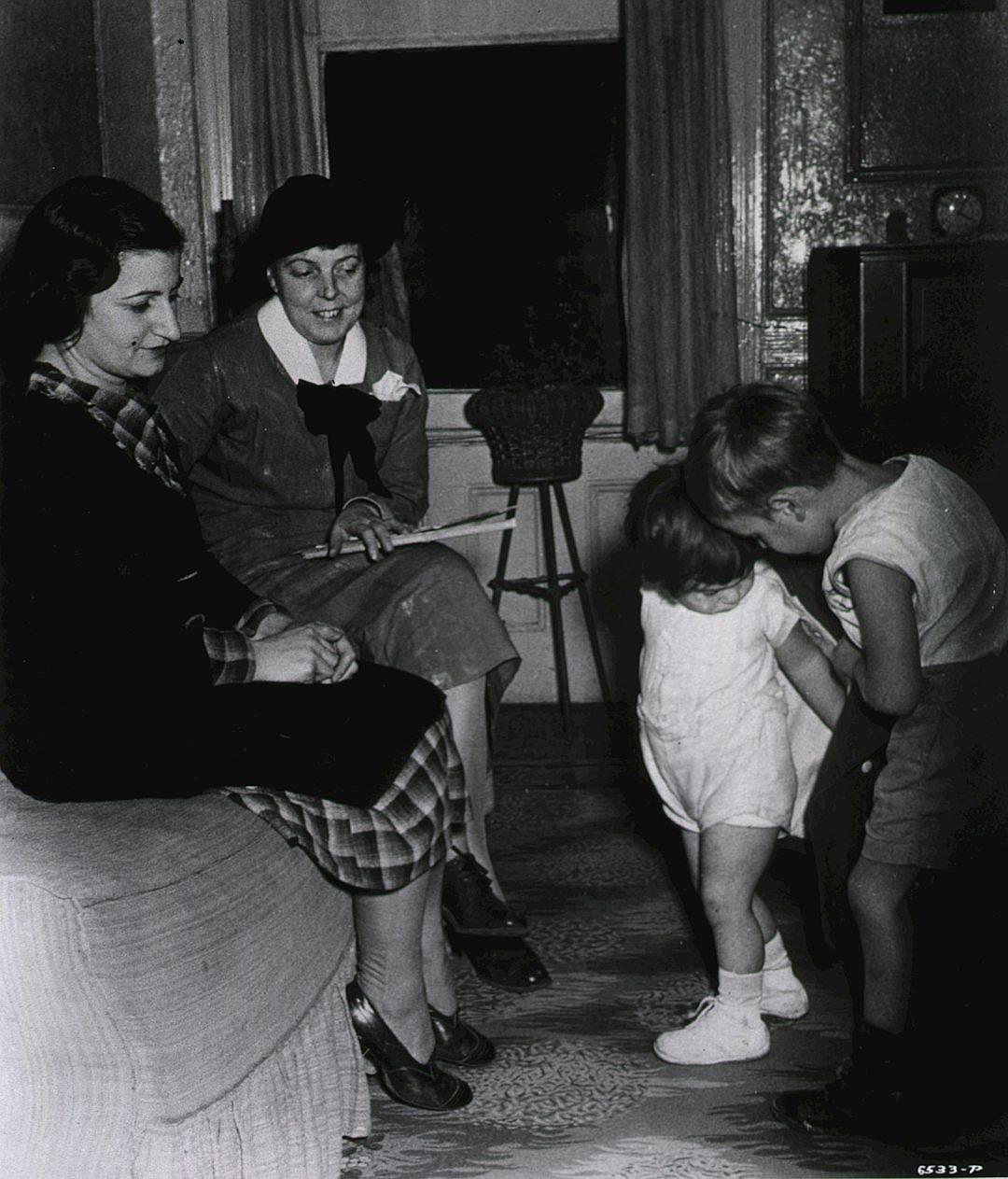 An old black and white photo shows two women sitting on a sofa, one wearing a plaid skirt and shoes while looking at another woman standing in front with her hands folded across her chest. She wears a plain short sleeve top, long pants, and child-like ankle socks. They look down at a small girl playing near them. The room feels old fashioned and dark but there is light coming from the window. The little girl’s face peeks out behind a table. The mother looks happy, perhaps just having seen it.