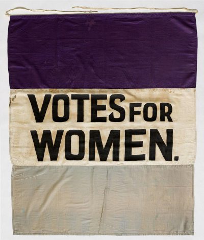 A banner with the words "VOTE FOR WOMEN" on it, purple and white cloth, designed in the style of Edward Gingerlach in collaboration with", "ROdata / OOTR, painted with black ink letters. The flag is hung at an angle, with three horizontal sections of light gray fabric, dark cream-colored linen, and deep lavender velvet. It's displayed against a plain background, showcasing its detailed construction and vibrant colors.