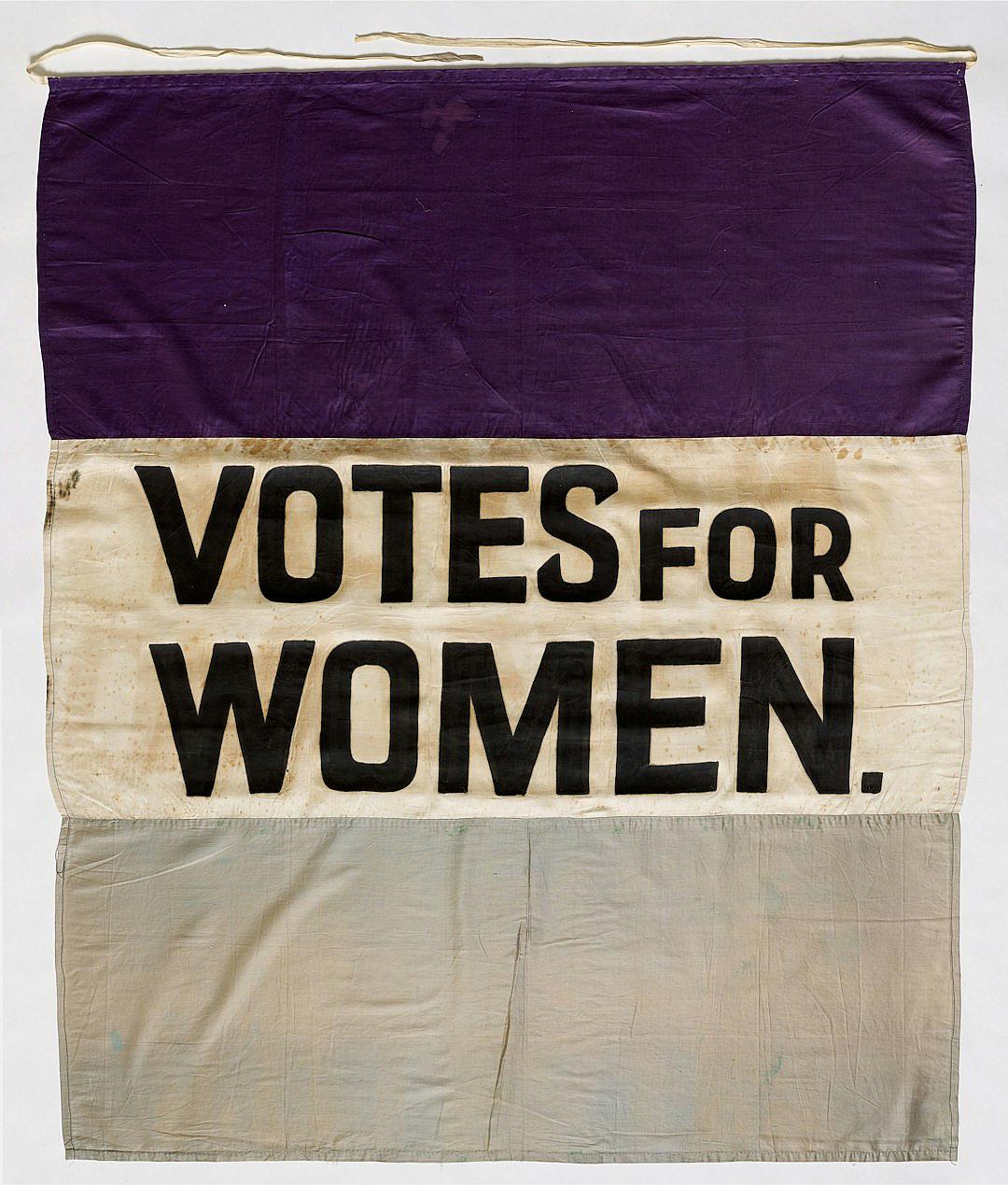 A banner with the words “VOTE FOR WOMEN” on it, purple and white cloth, designed in the style of Edward Gingerlach in collaboration with”, “ROdata / OOTR, painted with black ink letters. The flag is hung at an angle, with three horizontal sections of light gray fabric, dark cream-colored linen, and deep lavender velvet. It’s displayed against a plain background, showcasing its detailed construction and vibrant colors.