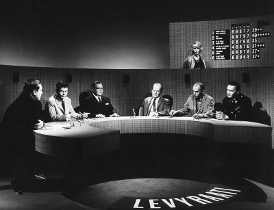 a black and white photograph of the first season panel from " Liverpool City" on British TV, with five guests sitting around an oval shaped table in front of camera lights, they look friendly but serious as if it is a game show, one host sits at head table facing all three other europeanstyle male visitors wearing suits and two female Coffeinastyle artists. The studio wall behind them has large numbers showing over night's intweather data, with text reading 'Martine Stella' written below in big letters. In the style of documentary photography.