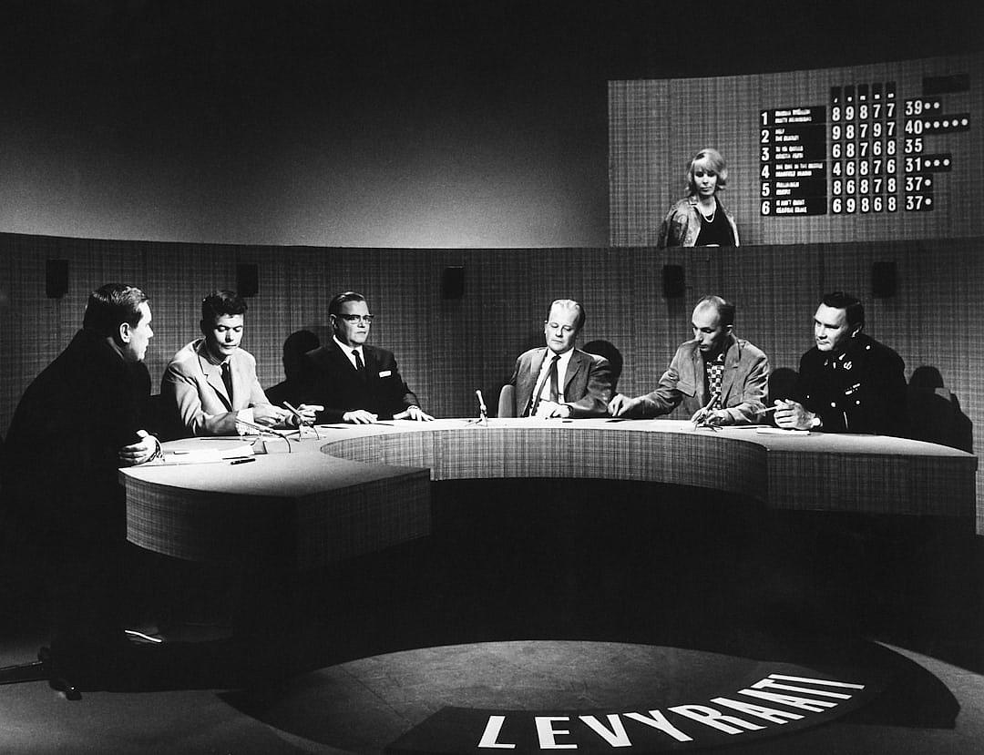 a black and white photograph of the first season panel from ” Liverpool City” on British TV, with five guests sitting around an oval shaped table in front of camera lights, they look friendly but serious as if it is a game show, one host sits at head table facing all three other europeanstyle male visitors wearing suits and two female Coffeinastyle artists. The studio wall behind them has large numbers showing over night’s intweather data, with text reading ‘Martine Stella’ written below in big letters. In the style of documentary photography.