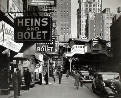 A black and white photograph of the streets in New York City from around the 1930s, with people walking on both sides, buildings in the background, street signs featuring large letters reading "HEINZ AND bolet" in the style of B的一员.