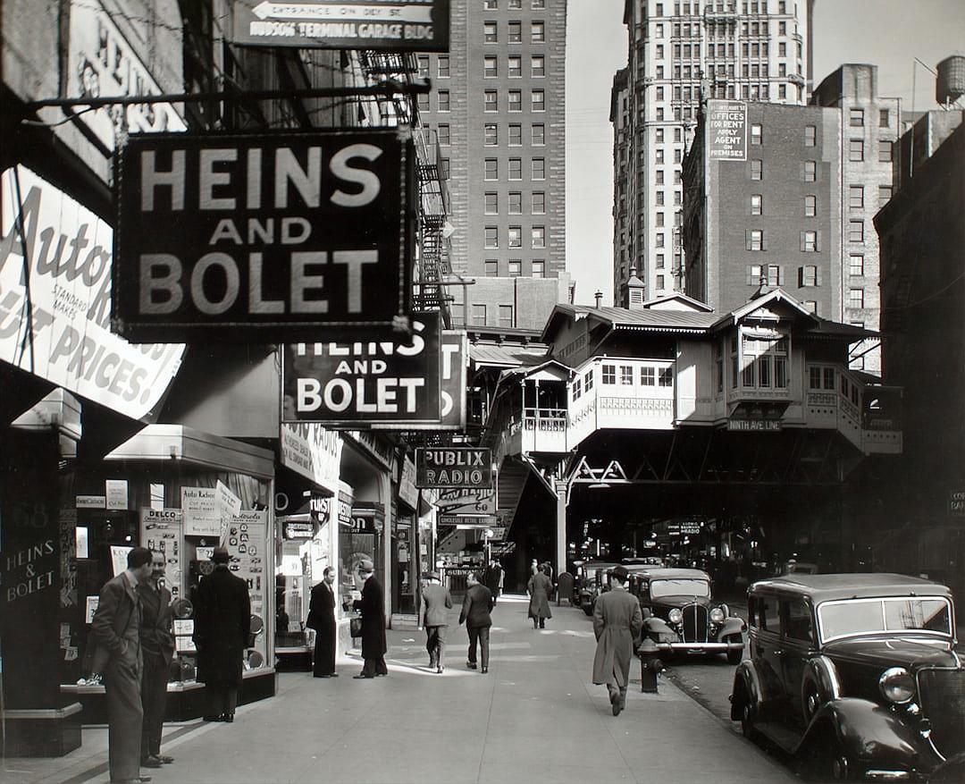 A black and white photograph of the streets in New York City from around the 1930s, with people walking on both sides, buildings in the background, street signs featuring large letters reading “HEINZ AND bolet” in the style of B的一员.