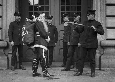 black and white photo of police officers in uniform, holding batons while looking at santa claus who is wearing his traditional suit with the word "LA" on it. They stand outside an old building. Santa has long hair and beard. The location appears to be San Francisco's city hall. One officer stands next to him pointing his hand toward us. It looks like they have just caught santa. The photo appears to be from the 1920s in the style of an old photograph from that era with long hair and beard on santa claus.