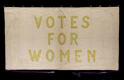 A banner with the words "VOTE FOR WOMEN" embroidered in yellow on white linen, with a black background and purple string for hanging, in the style of a vintage photograph with golden age aesthetics.