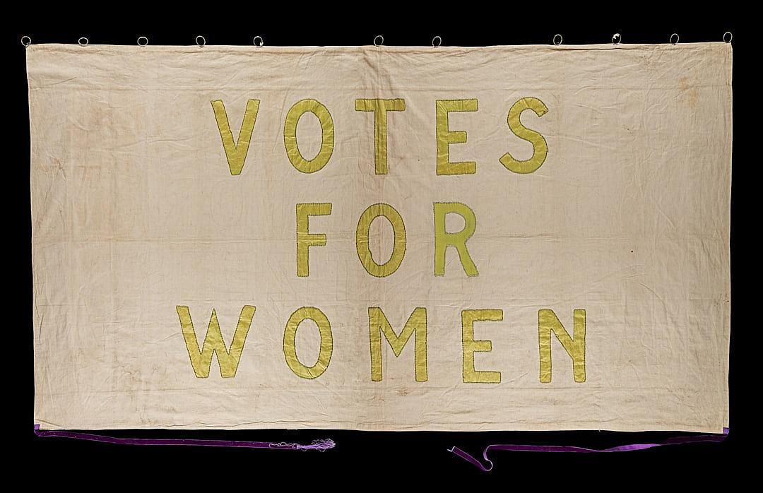 A banner with the words “VOTE FOR WOMEN” embroidered in yellow on white linen, with a black background and purple string for hanging, in the style of a vintage photograph with golden age aesthetics.