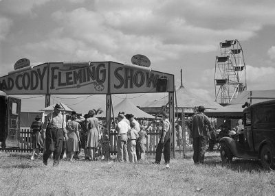 Black and white photo of the "Cody LaneAnthony wi dice show" at an old carnival in rural Texas, with people gathered around to watch the show, vintage cars parked nearby, a Ferris wheel visible behind them, and clouds overhead, capturing a sense of excitement and community during mid20th century American life.,