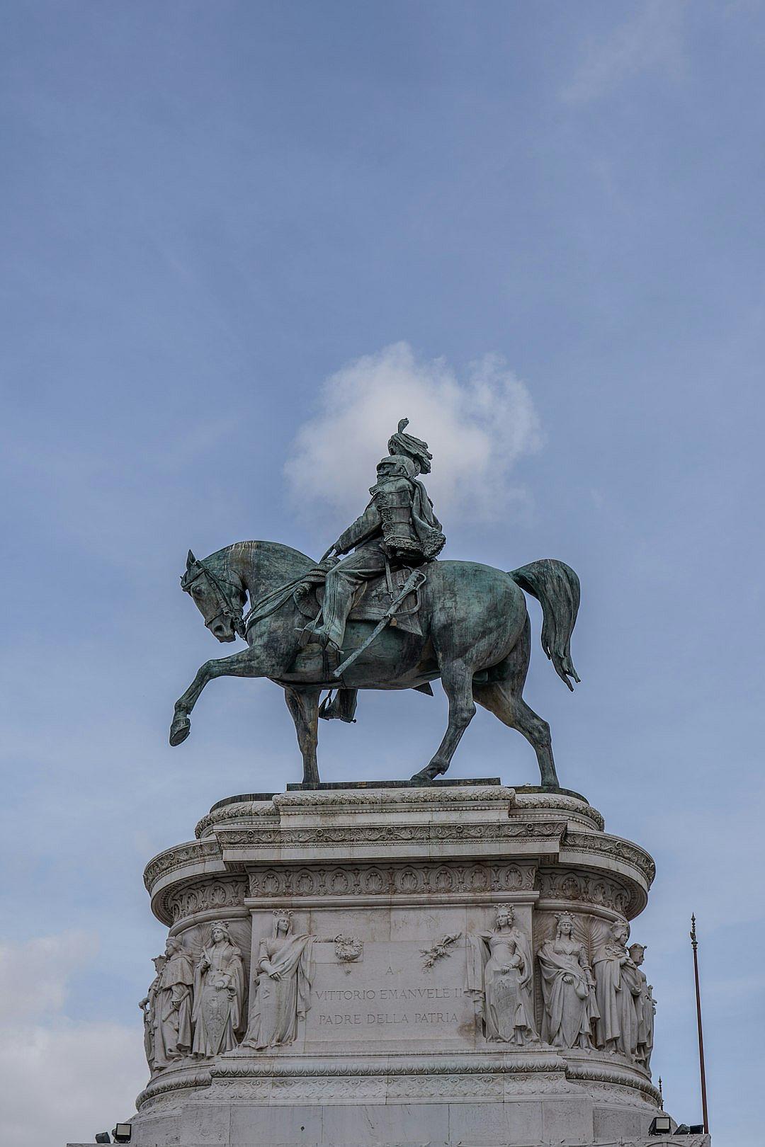 A large bronze statue of the general in battle on horseback, sitting atop an elegant pedestal with classical motifs and surrounded by statues depicting historical events from Italian history. The background is a blue sky. The photo was taken with a Sony A7R IV in the style of documentary photography.