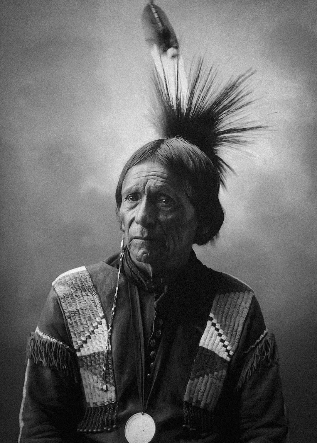 black and white photograph of an old Native American man wearing traditional , with hair made up in black feathers on top his head, he is facing forward looking at the camera