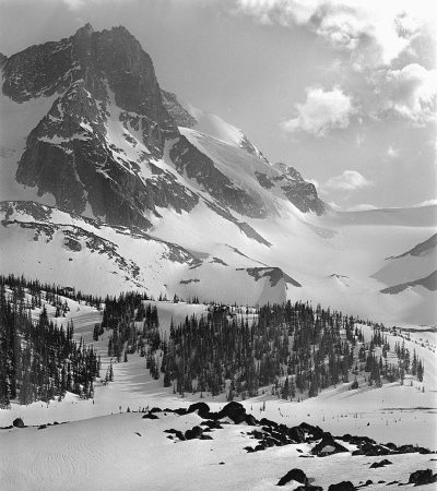 Black and white photograph of the snow-covered peaks of British Columbia's Elaffel Mountain, with skiers gracefully gliding down its slopes in the style of mid-20th century photography. The surrounding landscape is blanketed in pristine snow. In sharp focus against the backdrop of distant mountains and forests, there is an aura of tranquility as if time has come to stand still for just one moment. It is a wide shot captured with an ultra realistic style of photography.