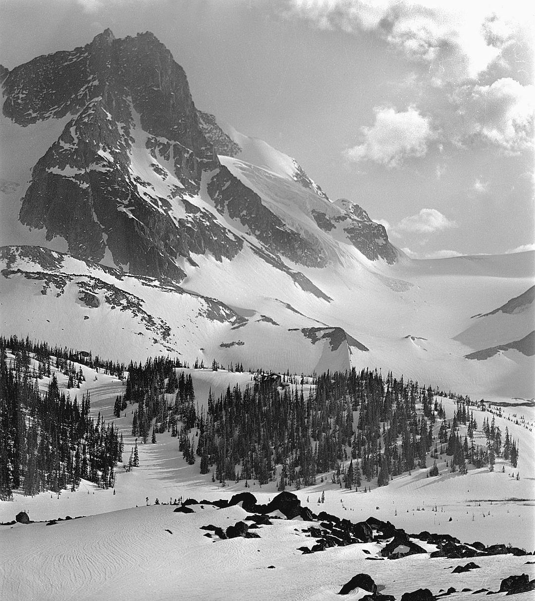 Black and white photograph of the snow-covered peaks of British Columbia’s Elaffel Mountain, with skiers gracefully gliding down its slopes in the style of mid-20th century photography. The surrounding landscape is blanketed in pristine snow. In sharp focus against the backdrop of distant mountains and forests, there is an aura of tranquility as if time has come to stand still for just one moment. It is a wide shot captured with an ultra realistic style of photography.