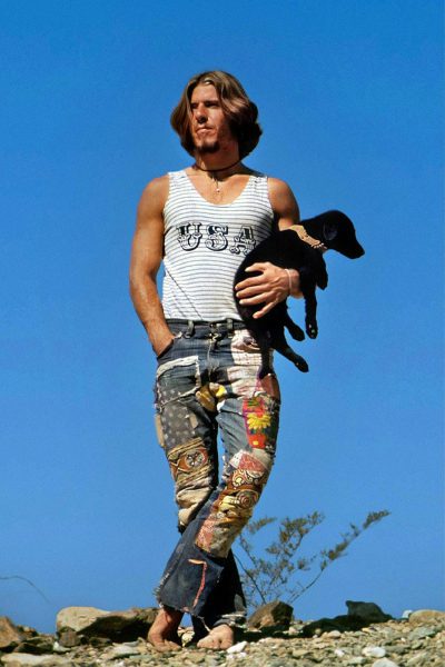 A photo of an athletic, longhaired man in jeans walking on the beach in California in summer with his black puppy. He is wearing a striped tank top that says "uteur". He has patchwork pants made from different fabrics. The sky behind him should be clear blue. Shot in the style of photographer [David LaChapelle](https://goo.gl/search?artist%20David%20LaChapelle).