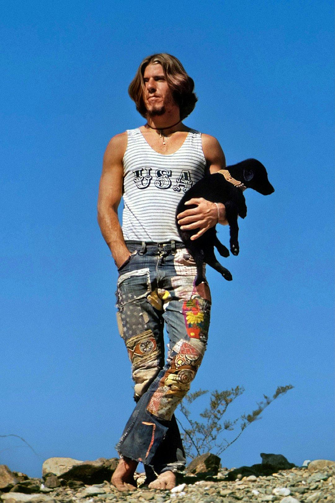 A photo of an athletic, longhaired man in jeans walking on the beach in California in summer with his black puppy. He is wearing a striped tank top that says “uteur”. He has patchwork pants made from different fabrics. The sky behind him should be clear blue. Shot in the style of photographer [David LaChapelle](https://goo.gl/search?artist%20David%20LaChapelle).