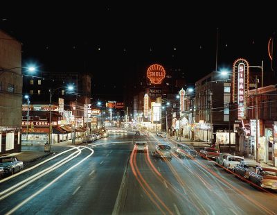 A night scene of downtownhouse, Canada in the late '50s and early '60s with neon signs glowing brightly on buildings along both sides of an empty street, cars passing by at high speed. The city's skyline is visible behind the bustling streets, with vibrant lights illuminating its architecture. A large billboard for "RUN timed agency" hangs above one building. This photo captures the lively atmosphere of urban life during that era.