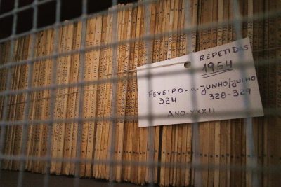 A stack of yellowed papers, each with "RE/name" and the date written in black ink. A white tag hangs from one paper that reads '4 JUN', with an arcade game number "329". The background is a wall covered entirely by wooden slats of different sizes, creating a dense pattern. There's some text on top right corner saying ‘ANOL’ and underneath below them is handwritten 'synthwave'. The style of the artwork appears to be in the style of Fenuissono de ferro.