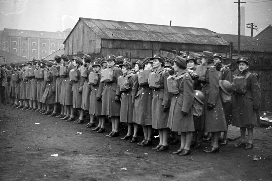 A group of women in work uniforms stand on the side of an industrial building, lined up and ready to go into action during World War II. They wear long coats over their dresses or pants with sleeves rolled down for protection against cold weather. Some have buttons at each shoulder line that could be used as backpack straps. The scene is captured from behind them in the style of photographer James K yothe.