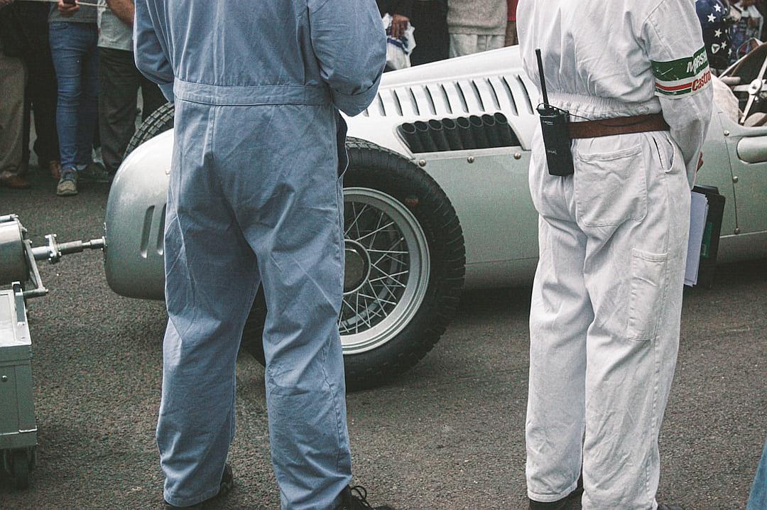 An old photograph of two men in blue overalls and white jumpsuits standing next to a silver sports car at the Goodwood Festival of Speed, vintage photography, 35mm film, grainy, film noise, in the style of Anonymous.