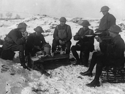 A black and white photograph of the first world war, soldiers sitting around eating lunch in snow covered field on a wooden bench with their arms crossed or holding food plates or mugs. They all wear uniforms with flat helmets that cover part of thier faces. The background is a snowy landscape. It's winter time and there is little light but they can see each other well enough to talk over dinner. One soldier has his legs propped up onto another. All the men look tired yet contented.