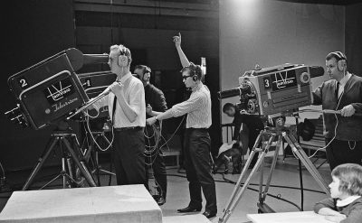 A behind the scenes photo of an early television set shows cameras and microphones. Two men in suits stand on stage next to each other holding their hands up high as if they were celebrating something. One man is blonde wearing headphones standing near a camera on the right side. Another person stands far away pointing his finger at him while talking into a microphone attached to his left ear. People sit around watching them from their seats. Children play nearby in the style of [Norman Rockwell](https://goo.gl/search?artist%20Norman%20Rockwell).