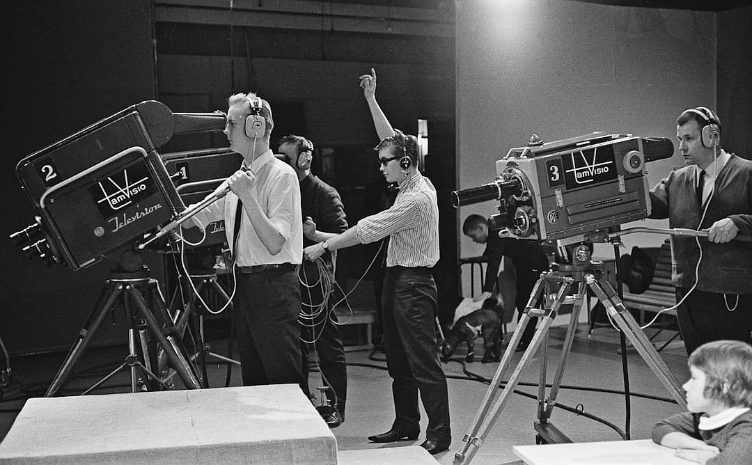 A behind the scenes photo of an early television set shows cameras and microphones. Two men in suits stand on stage next to each other holding their hands up high as if they were celebrating something. One man is blonde wearing headphones standing near a camera on the right side. Another person stands far away pointing his finger at him while talking into a microphone attached to his left ear. People sit around watching them from their seats. Children play nearby in the style of [Norman Rockwell](https://goo.gl/search?artist%20Norman%20Rockwell).