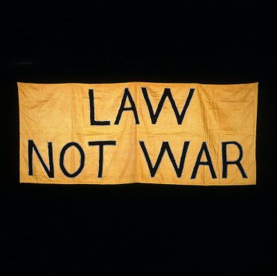 A yellow banner with the words "Law, not War" written in black letters on it. The background is dark and simple. It appears to be made of fabric or cloth. There's no other text visible on the flag except for these two lines. In its center lies an emblem that could represent peace or war, but without any clear details beingrays. This symbolizes a message about battling fire while maintaining balance between lawful forces and open battle, possibly as part of military selfportrait photography techniques.