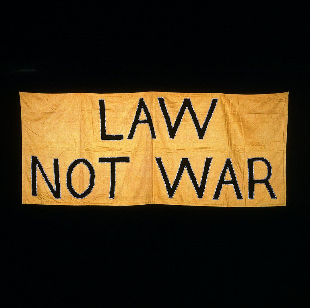 A yellow banner with the words “Law, not War” written in black letters on it. The background is dark and simple. It appears to be made of fabric or cloth. There’s no other text visible on the flag except for these two lines. In its center lies an emblem that could represent peace or war, but without any clear details beingrays. This symbolizes a message about battling fire while maintaining balance between lawful forces and open battle, possibly as part of military selfportrait photography techniques.