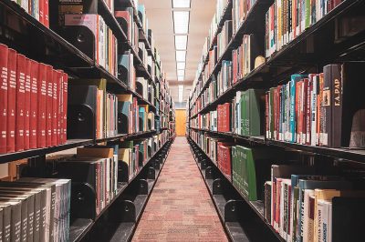 A library with rows of bookshelves filled with various books, shot from the perspective looking down at an empty path between two sets of shelves. The focus is on one side where colorful and black items stand out among others, creating contrast in color. There is light coming through from above, highlighting details like buttons or sleek surfaces on some equipment. This scene conveys calmness and order within its urban environment.