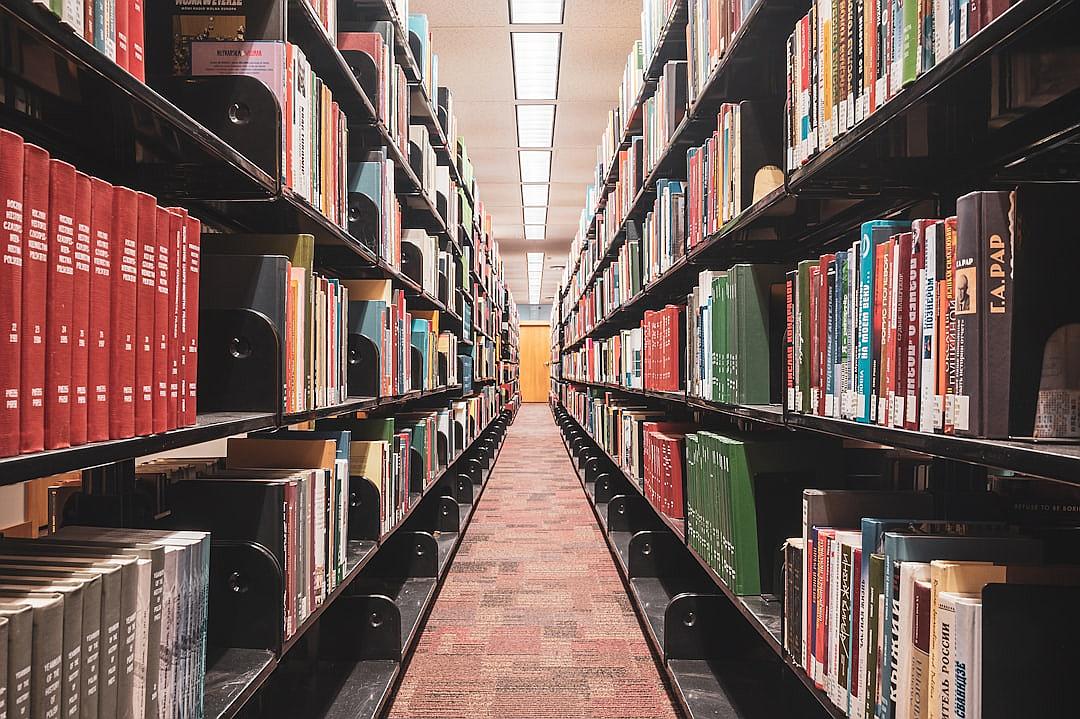 A library with rows of bookshelves filled with various books, shot from the perspective looking down at an empty path between two sets of shelves. The focus is on one side where colorful and black items stand out among others, creating contrast in color. There is light coming through from above, highlighting details like buttons or sleek surfaces on some equipment. This scene conveys calmness and order within its urban environment.