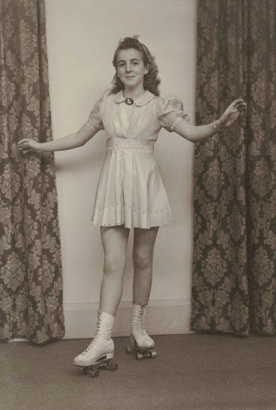 old photo, vintage, retro, girl on roller skates in short dress with puffy sleeves and white shoes smiling at the camera, standing, hands outstretched to her sides, arms straight above head level, standing, legs slightly bent at knees, full body portrait, front view, curtains behind woman