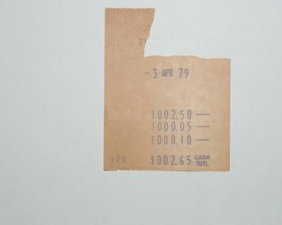 A small, crumpled beige cash register paper with the date "3 perennial apr'79" at top and numbers showing money amount in middle on white background. The title is "Six tight shops", a line of text under it reads" $025 CASHConnor T uno's". There’s an inscription "Azgrul O Robbie" on bottom right corner. . A photo taken by canon eos r6, wide angle lens,