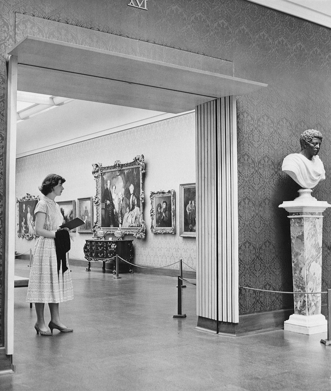 The National Geographic photo of an art gallery showed a woman standing next to a wall with classical paintings on the walls and marble statues in the corner, it was a black & white photograph from the 1950s.