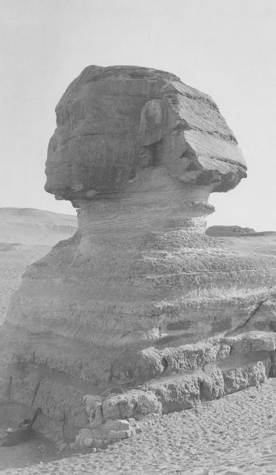 Black and white photograph of the sphinx, showing its profile in an open desert landscape. The sculpture is carved from stone with detailed features such as eyes, nose, mouth, ears, head shape, hair, body, tail, back and feet. There's sand around it and some small rocks nearby. In front there’s an empty space without any objects or people. It has a strong contrast between light and shadow, highlighting textures like roughness on different parts of his face and neck.