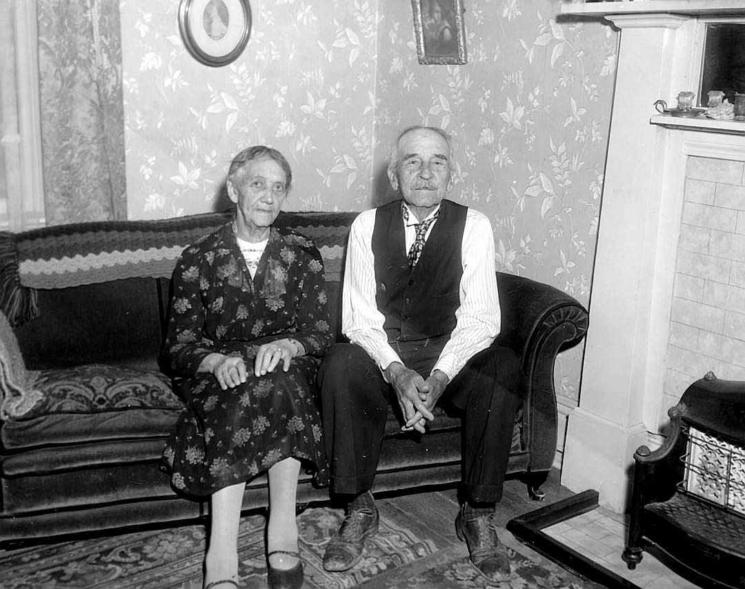 An old black and white photograph shows an elderly couple sitting on the couch in their living room. They look happy to be together. The woman is wearing a patterned dress with shoes and her hair is combed back. He wears slacks, a tie, a vest and dirty boots. There is wallpaper behind them with small floral patterns. There is a fireplace in front of it with a woodburning stove.