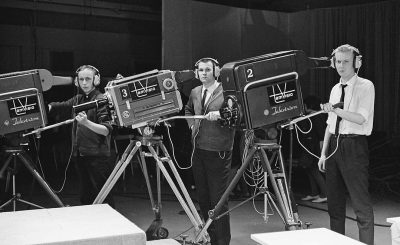 In the early days of television, in an empty studio with cameras and lights set up for live television, three young men stand behind their equipment dressed as news paparazzi wearing black suits and white shirts tied at the neck with navy blue ties. They hold microphones that have "TV melreeze" written on them. The atmosphere is excited but professional. Black & White film photography, shot in the style of ARRIFLEX35 BL Camera Canon K207 Prime Lens Full Shot, hyper realistic, detailed skin, natural lighting, HDR.