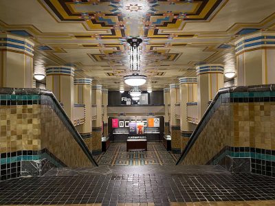 The interior of an Art Deco theater lobby with geometric tiling and colorful painted accents, two stairs lead to an axonometric film store counter above the carpeted floor, in the style of Art Nouveau with ornate light fixtures hanging from the ceiling, architectural photography.