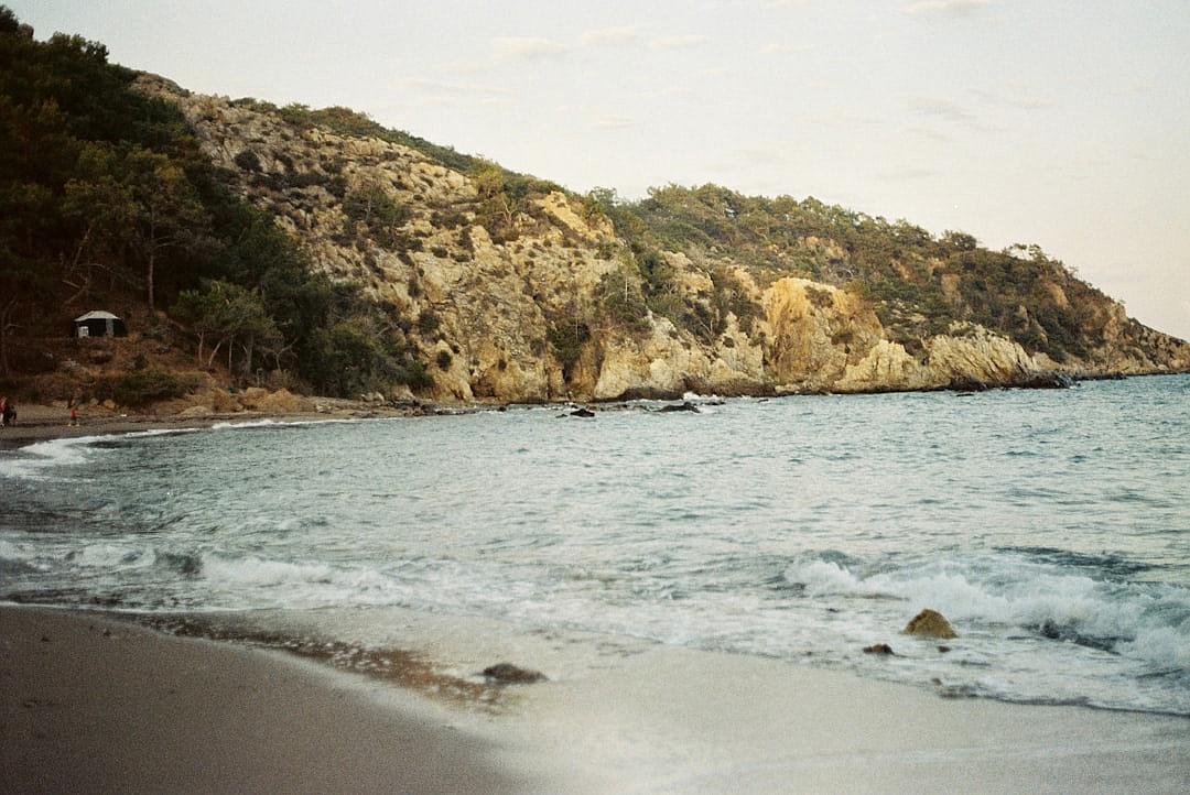 A beach in Greece, near the water is an area of rugged cliffs with trees and rocks, shot on Kodak Gold film, natural lighting, cinematic, grainy, analog photography in the style of analog photography.