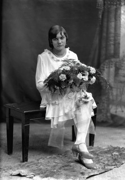 A black and white studio portrait of a young woman sitting in a chair holding a large bouquet with ribbons, wearing a white long sleeve blouse and pants from the 1920s. She has long hair in a short bob cut hairstyle and vintage shoes. It is a full body shot of her in a standing pose with a front view against a monochrome background, in the style of that era.