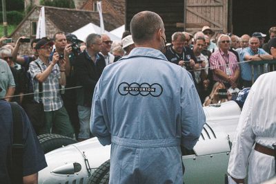 A man in an Audi jumpsuit stands with his back to the camera. On one side of him is an old white car, and behind it there are people taking pictures and standing around at a classic cars show in the English countryside. "Engineering creed" is written across the chest area of his shirt. A crowd watches a race between two vintage military vehicles in the style of a Pokémon show.