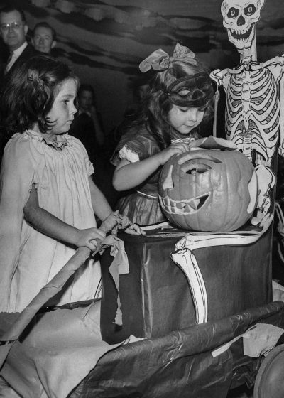 Vintage style, black and white photo of two girls carving pumpkins at an old-fashioned Halloween party in the family home with jack-o'-lanterns on boxes, surrounded by spooky decorations like skeleton toads, ghostly scenery, children's costumes and candy bags. The atmosphere is filled with excitement as they create their unique designs in the style of vintage Halloween.