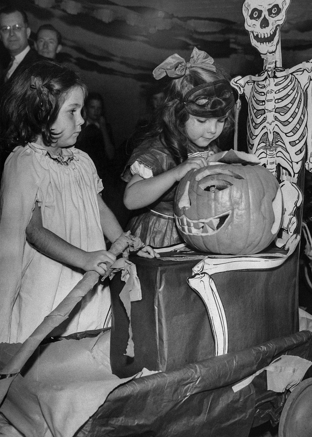 Vintage style, black and white photo of two girls carving pumpkins at an old-fashioned Halloween party in the family home with jack-o’-lanterns on boxes, surrounded by spooky decorations like skeleton toads, ghostly scenery, children’s costumes and candy bags. The atmosphere is filled with excitement as they create their unique designs in the style of vintage Halloween.