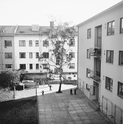 A black and white photo of the outside courtyard area at a school in Sweden. There is one tree with children playing on it. You can see some people walking around between buildings. There is an apartment building to the right side. The photo was shot from above with 35mm film grain in the style of a documentary photograph.