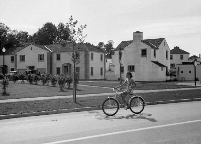 A black and white photo of an American suburban neighborhood in the mid 20th century, with two story houses on one side of the street. A woman is riding her bike down the middle of the street as she rides past each house. The style should be reminiscent of classic film photography from that era. Use a Hasselblad camera to capture sharp details and natural colors. Make sure to focus on capturing intricate textures of skin, hair,  and surroundings. It must look like a real photograph taken in the style of [Man Ray](https://goo.gl/search?artist%20Man%20Ray) or [Ansel Adams](https://goo.gl/search?artist%20Ansel%20Adams).